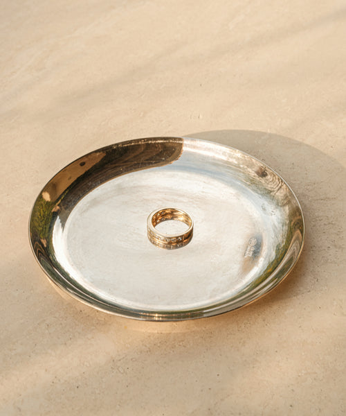 The Stevie Grey Diamond Band, a fine jewelry gold ring with delicate engravings, rests on a reflective silver plate atop a beige surface, bathed in soft natural light.