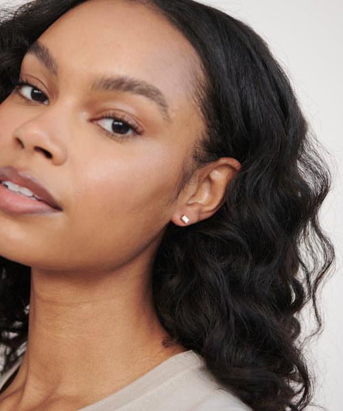 A woman with wavy dark hair and smooth skin wears Ivy Emerald Cut Studs, elegant 14k gold earrings. She is shown in close-up, gently tilting her head and gazing softly at the camera.