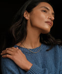A woman wearing a blue knit sweater, gold necklaces, a ring, and Ivy Emerald Cut Studs in 14k gold looks to the side with her hand on her shoulder against a dark background.