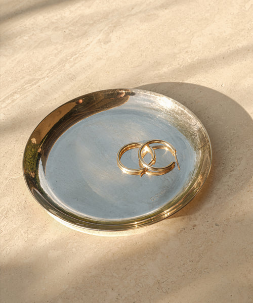 A pair of Medium Eloise Hoops in 14k gold rests on a round, metallic tray with a gold rim. The tray sits atop a beige surface, where gentle sunlight and soft shadows highlight the timeless elegance of heirloom jewelry.
