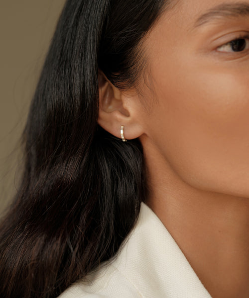A close-up of a persons ear wearing Merit Baguette Hoops—elegant 14k gold earrings with tiny embedded stones. Their long dark hair and beige top complement the timeless beauty of this heirloom jewelry against a neutral, blurred background.