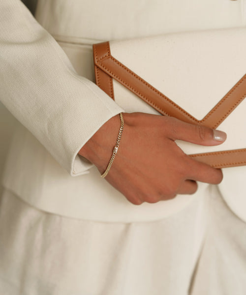 A person in a cream-colored suit holds a matching clutch and accessorizes with fine jewelry, featuring the Merit Emerald Cut Bracelet on their wrist and neatly manicured nails.