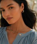A woman wearing a light blue blouse, gold hoop earrings, and the Merit Emerald Cut Necklace gazes softly at the camera outdoors in natural light.