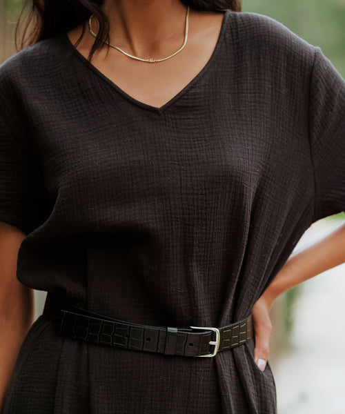 A person wears a textured black V-neck short-sleeve dress with a black belt featuring a silver buckle, accessorized with the Merit Emerald Cut Necklace.
