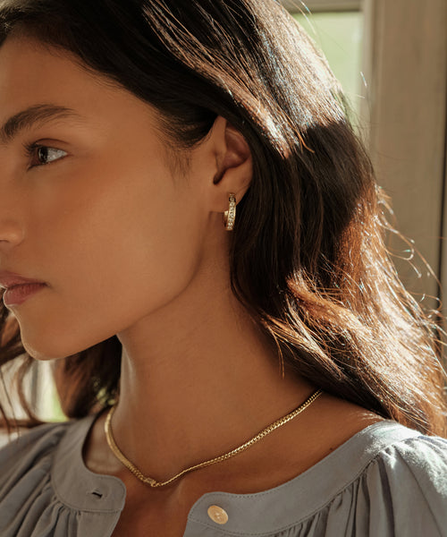 A woman with long brown hair wears Stevie Grey Diamond Hoops and a gold necklace, dressed in a light gray buttoned blouse. She stands in soft natural light, showing only the left side of her face and neck.