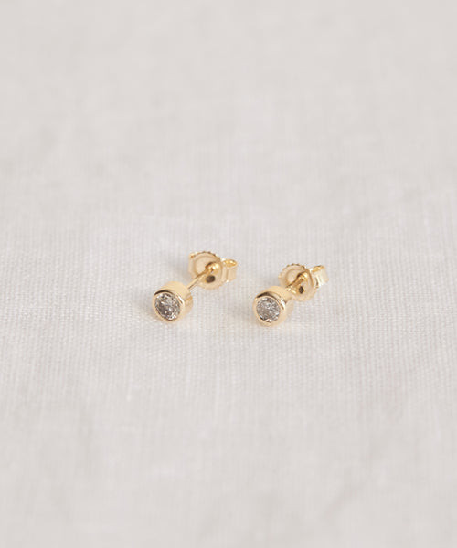 A pair of Stevie Grey Diamond Studs in 14k gold, featuring small round grey diamonds, displayed on a light, textured fabric background.