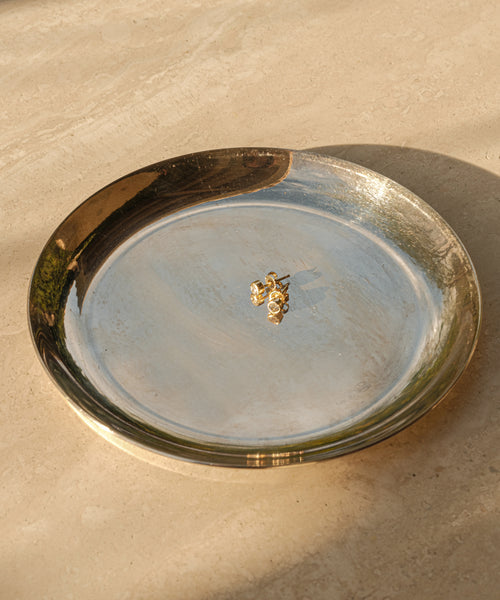 A pair of Stevie Grey Diamond Studs in 14k gold rest on a shiny round metallic plate atop a beige stone surface, softly lit—an elegant and timeless choice for any Jenni Kayne jewelry collection.