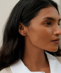 A woman with long dark hair wears a white collared shirt and the Stevie Grey Diamond Studs, gazing thoughtfully to the side against a softly blurred neutral background.