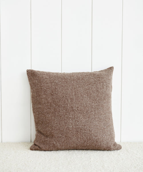 An Alpaca Basketweave Pillow in a square shape and brown color is propped upright against a white paneled wall, resting on a light-colored carpet.