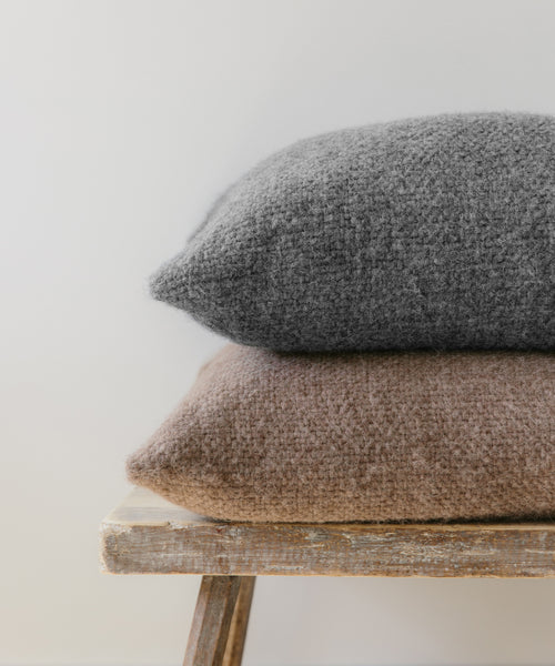 A gray Alpaca Basketweave Pillow and a brown cushion are stacked on a rustic wooden bench against a plain light-colored background.