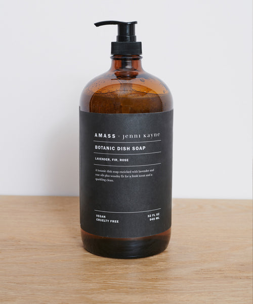 A brown reusable glass bottle labeled AMASS x Jenni Kayne Dish Soap rests on light wood, featuring lavender, fir, and rose scents. The vegan formula is cruelty-free.