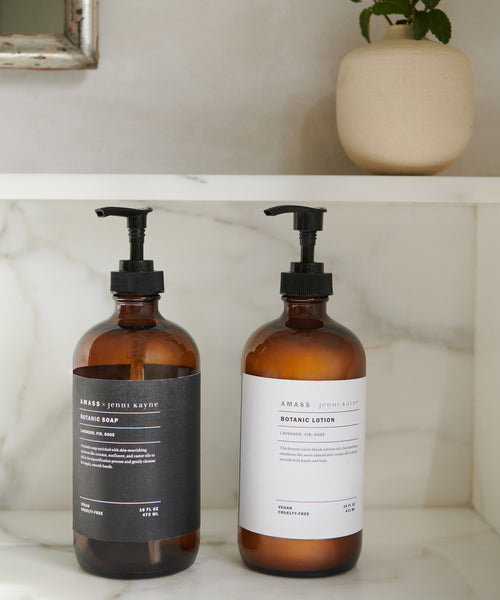An amber glass bottle labeled AMASS x Jenni Kayne Dish Soap sits elegantly on a white marble shelf, its reusable design highlighted by a beige vase with a small green plant adding freshness in the background.