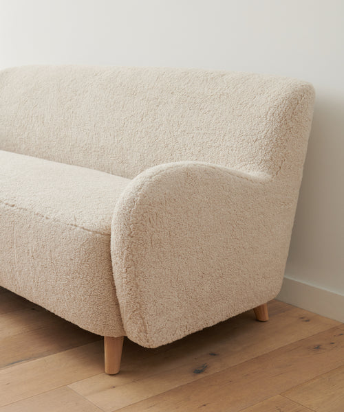 The Aspen Sofa with rounded armrests and wooden legs sits on a light wood floor against a plain white wall.