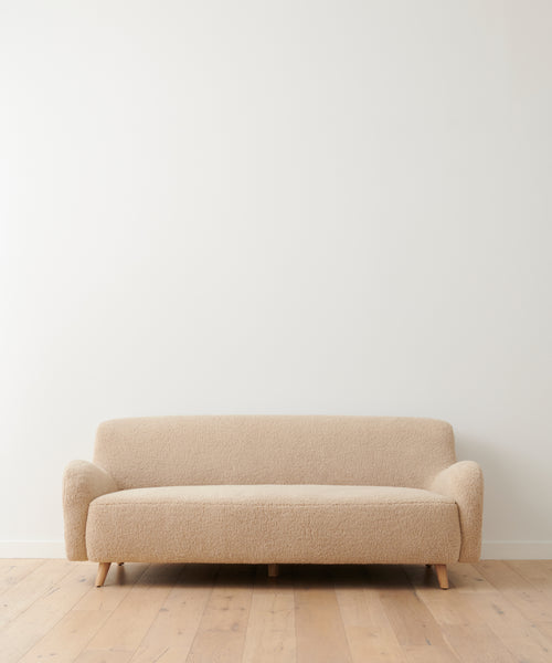 A beige Aspen Sofa with textured fabric and wooden legs stands against a white wall on a light wood floor in a minimalist modern room.