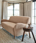 A sofa upholstered in Fabric Swatch Oatmeal Sherpa sits on a textured rug in a bright room with large windows and cream curtains, paired with a rustic wooden side table holding a candle and dried flowers.
