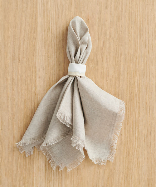 A beige linen napkin with frayed edges is folded and held by a Ceramic Napkin Rings Set of 4, handmade in California, and placed on a light wooden surface.