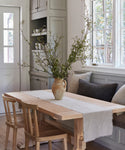 A light wood dining table with two chairs and a bench sits in a cozy kitchen nook. A ceramic vase with tall branches is centered on the table, which is styled with the Frayed Linen Runner. Built-in shelves and large windows fill the background.