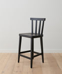 The Hampton Counter Stool, a simple black oak Windsor-style chair with a slatted backrest and four legs, is set on a light wooden floor against a plain white wall.