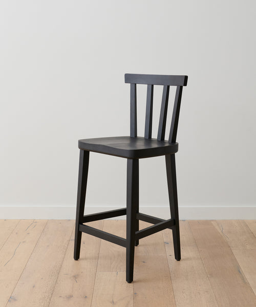 The Hampton Counter Stool, a simple black oak Windsor-style chair with a slatted backrest and four legs, is set on a light wooden floor against a plain white wall.