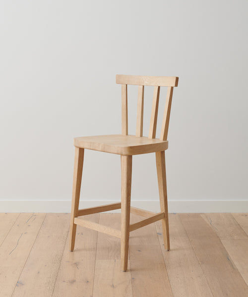 The Hampton Counter Stool, crafted from White Oak with a slatted backrest and four legs, sits on a light wooden floor against a plain white wall.