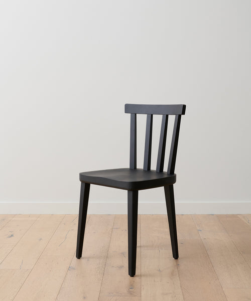 The Hampton Dining Chair in black oak, with three vertical backrest slats and a minimalist design, stands on a light wooden floor before a plain white wall.