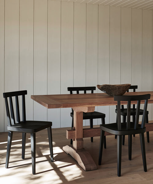 A minimal dining area features a wooden table with four black Hampton Dining Chairs and a rustic wooden bowl, set against light-paneled walls and a wood floor. Sunlight casts gentle shadows across the inviting space.