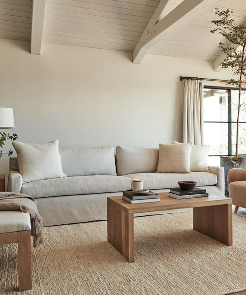A modern living room features a Harbor Sofa in natural linen, two beige pillows, a wooden coffee table with books and a candle, a soft rug, a floor lamp, and beige curtains letting natural light in.