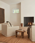 A cozy living room featuring two Harbor Chairs in natural linen, a wooden coffee table with books, a brown rug, a fireplace with a warm fire, a ceramic vase, and natural light streaming through a small window.
