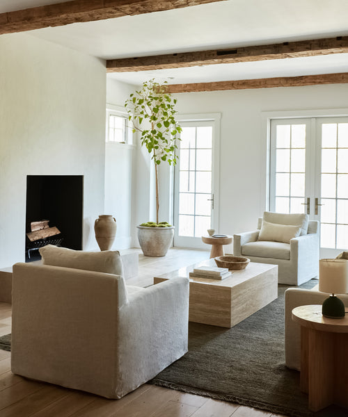 A modern living room showcases Harbor Chairs in natural linen, a wooden coffee table, ceramic vases, a potted plant, exposed wood beams, and large windows that fill the space with light.