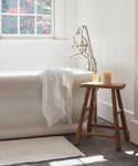 A white bathtub stands near a window with a towel draped over the edge and sunlight brightening the neutral-toned bathroom. The Bath Mat in soft ivory lies beneath, while a wooden stool holds two candles.