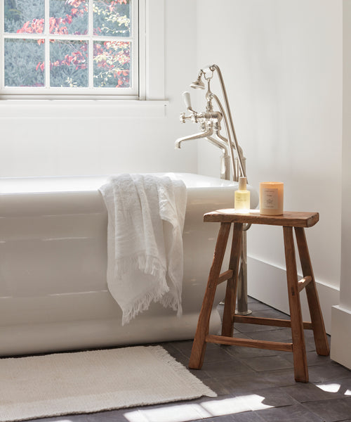 A white bathtub stands near a window with a towel draped over the edge and sunlight brightening the neutral-toned bathroom. The Bath Mat in soft ivory lies beneath, while a wooden stool holds two candles.