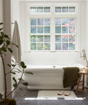 Natural light fills a bright bathroom featuring a white freestanding bathtub under large windows, an ivory bath mat (Bath Mat) beside the tub, a towel on the wall, green plant, and candle on the tub’s edge.