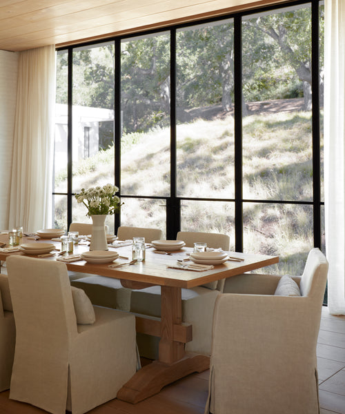 A modern dining room includes a wooden table set for six, each seat featuring the Sunset Dining Arm Chair Slipcover in sandstone linen. A vase with flowers sits on the table, and floor-to-ceiling windows showcase a sunlit grassy hillside.