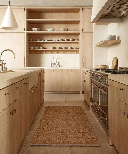 Modern kitchen with light wood cabinets, open shelves, brass fixtures, two sinks, and a large stove. The Bramble Rug in natural jute sits on a beige tiled floor, complementing the clean, minimalist design. Dishes and bowls are neatly stacked on display.