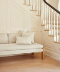 The Ynez Daybed in light beige Belgian linen, featuring two matching cushions, is placed by a wooden staircase with white spindles and natural steps in a bright, minimalist room with paneled walls.