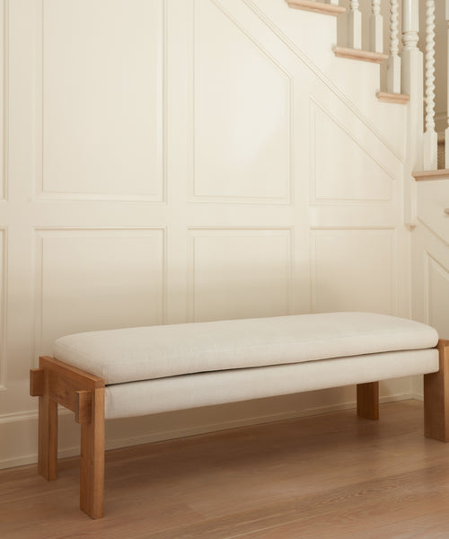 The Ynez Bench, crafted from Belgian Libeco linen with a light cushion, rests against a cream-paneled wall by a staircase with white spindles and wood railings. Light wood floors complete the serene, luxurious entryway look.