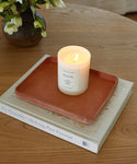 A lit white candle in a glass jar sits on the Leather Catchall, handmade in Los Angeles, which rests on two stacked books on a wooden table. In the background, a vase with white flowers is visible.