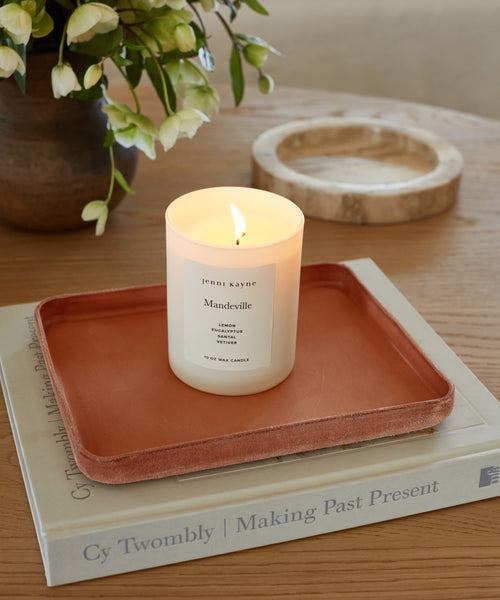 A white candle in a glass jar rests on the Leather Catchall, handmade in Los Angeles, atop an art book on a wooden table, with a vase of white flowers and a round beige dish visible in the background.