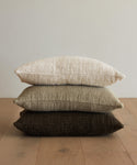 Three Luna Pillows—handwoven in ivory, light beige, and dark brown—are stacked neatly on a wooden floor against a plain light-colored wall.