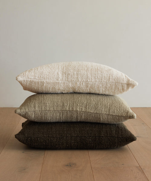 Three Luna Pillows—handwoven in ivory, light beige, and dark brown—are stacked neatly on a wooden floor against a plain light-colored wall.