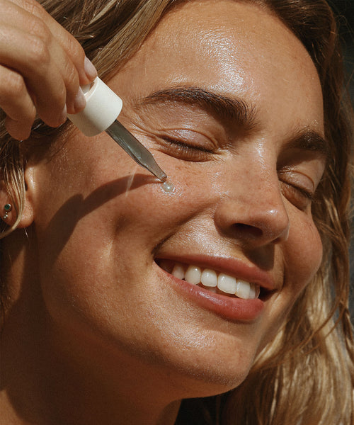 A person with radiant skin smiles with eyes closed as they apply a drop of The Reset Duo to their cheek, sunlight enhancing their healthy complexion and highlighting the benefits of this nourishing skincare routine.
