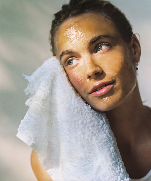 A woman with glowing, dewy skin gently presses a white towel to her face, her radiant complexion reflecting the nourishing benefits of The Reset Duo. Soft light highlights her features against a neutral background.