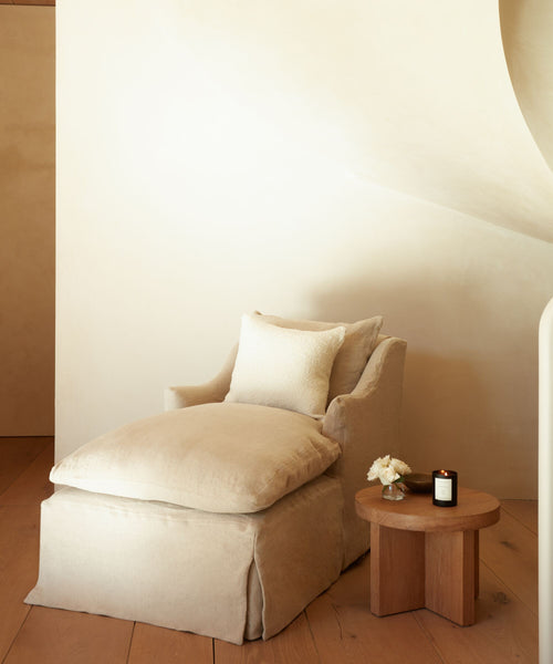 A cozy beige chaise lounge with an Alpaca Basketweave Pillow sits by a wooden side table with a lit candle and vase of white flowers, set against a light, minimalist background.