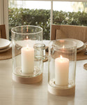 Two lit white pillar candles sit in Stowe Eco Glass Vases with light wooden bases, placed on a dining table set with plates, glasses, and woven placemats near a window.