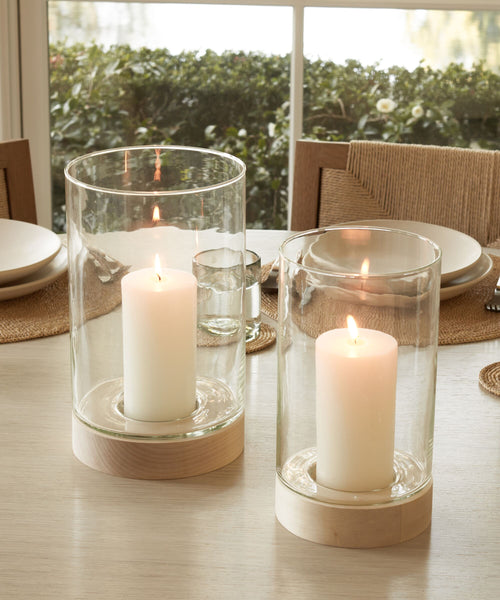 Two lit white pillar candles sit in Stowe Eco Glass Vases with light wooden bases, placed on a dining table set with plates, glasses, and woven placemats near a window.