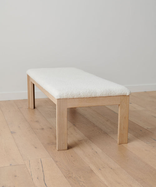 The Otto Bench features a minimalist white oak frame and ivory wool boucle cushion, shown on a light wood floor with a plain white backdrop.