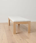 The Otto Bench, handcrafted from white oak, features a natural linen cushion in light tones. Shown on a light wood floor with a plain white backdrop.