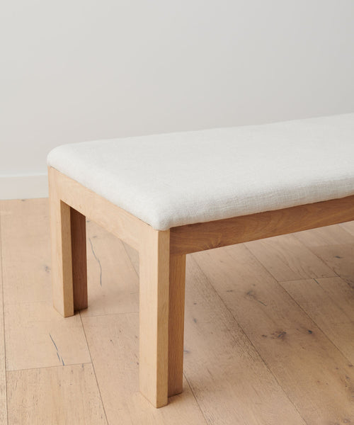 The Otto Bench, made from handmade white oak and topped with a natural linen cushion, sits on a light wood floor by a plain white wall. The image shows only one end of the bench.