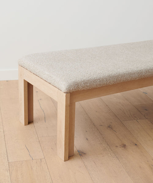The Otto Bench is a minimalist piece crafted from white oak with a light beige upholstered seat, placed on a pale wood floor against a plain white wall.