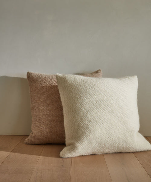 A cream Alpaca Basketweave Pillow sits on a light wooden floor in front of a larger beige pillow, both resting against a plain gray wall.